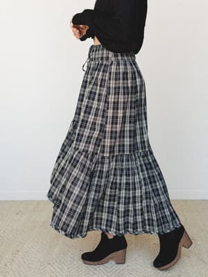 A tiered plaid maxi skirt in black and cream styled with a black long-sleeve top and chunky suede ankle boots — the kind of outfit that makes a Tuesday feel intentional.