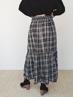 A plaid tiered maxi skirt in black and cream is shown from the back, styled with a black long-sleeve top and black block-heel ankle boots — easy, grounded, and the kind of outfit that works harder than it looks.