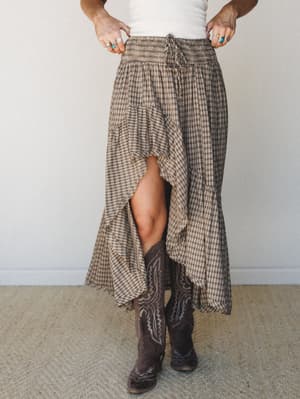 A flowy plaid high-low maxi skirt in warm chocolate tones styled with dark cowboy boots and a white crop top — the kind of outfit that makes a farmers market feel like a main character moment.
