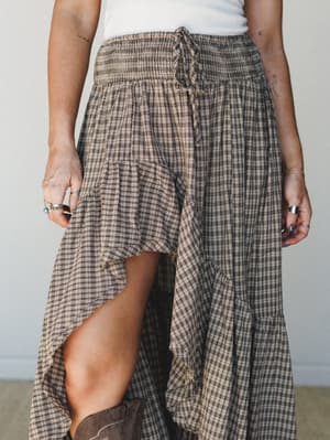 A close-up of a flowy plaid maxi skirt with a smocked waistband and high-low hem, styled with a white tank and brown boots — earthy, relaxed, and effortlessly lived-in.