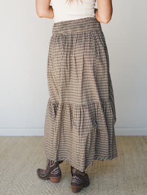 Shot from behind, a tiered plaid maxi skirt in warm chocolate and tan pairs with a simple white tee and dark brown cowboy boots — the kind of effortless fall outfit that feels equal parts cozy and intentional.