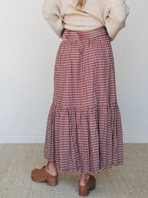 Shot from behind, a tiered plaid midi skirt in deep red and cream pairs effortlessly with a chunky cream knit sweater and wood-studded clogs — the kind of autumn outfit that makes you want to walk through a farmer's market.