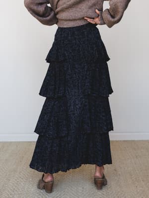 Shot from behind, a model in a cozy taupe sweater and block-heel mules wears a dramatic tiered maxi skirt in a dark navy and black abstract print — the kind of outfit that feels equally at home at a dinner out or a Sunday farmer's market.