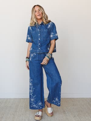 A matching embroidered denim set — cropped button-up shirt and wide-leg pants — covered in white floral vine embroidery that gives the whole look a vintage-boho feeling, styled with espadrille platform sandals and stacked beaded bracelets.