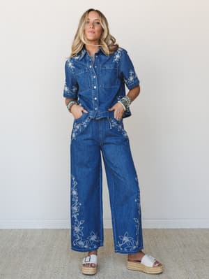 A full denim matching set — embroidered button-up shirt and wide-leg jeans — styled with stacked turquoise bracelets and white platform sandals against a clean neutral wall; the kind of outfit that does all the talking.