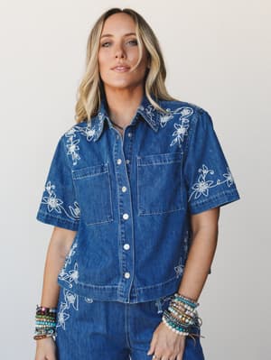 A cropped denim button-up shirt covered in white floral embroidery, styled with matching denim bottoms and stacked beaded bracelets — the kind of outfit that feels intentional without trying too hard.