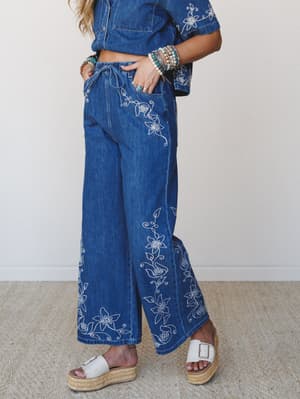 A medium-wash denim wide-leg pant covered in white floral embroidery pairs with a matching cropped denim top, styled with platform espadrille sandals and a stack of turquoise and pearl bracelets — the kind of outfit that feels effortless and a little