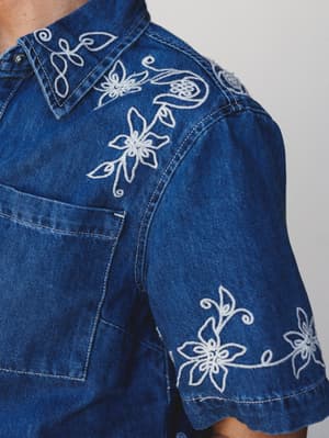 A close-up detail shot of a medium-wash denim top covered in delicate white floral and vine embroidery — the kind of piece that makes denim feel like something you'd actually get dressed up for.