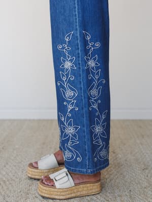 A close-up detail shot of wide-leg denim jeans with delicate white floral and vine embroidery running along the hem, paired with cream platform slide sandals on a woven rug — the kind of detail that makes you stop scrolling.