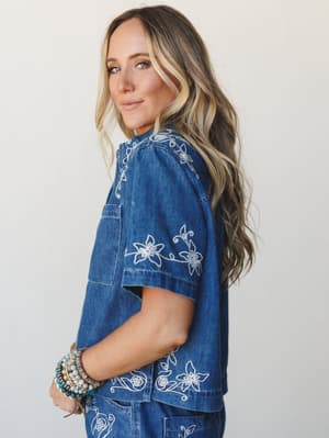 A relaxed denim set with white floral embroidery — worn with a stack of beaded bracelets and an easy side glance that feels effortlessly personal.