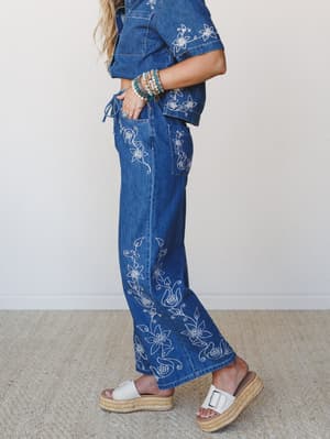 A side-profile shot of a denim matching set covered in delicate white floral embroidery — wide-leg pants and a cropped short-sleeve top styled with stacked turquoise bracelets and white espadrille platform sandals, shot clean against a soft white int