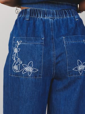 A close-up back view of medium-wash wide-leg denim pants with white floral embroidery on the patch pockets and an elastic waistband — the kind of detail you notice and can't stop thinking about.