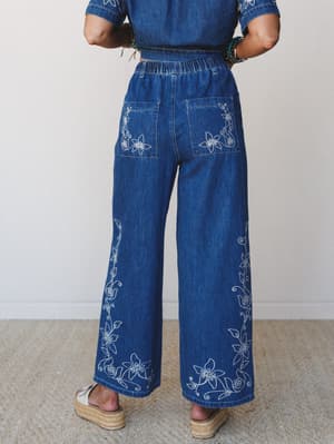 Shot from behind, a model wears wide-leg embroidered denim pants with a matching cropped denim top — the white floral embroidery trailing up the legs and across the back pockets is the whole personality of this outfit.