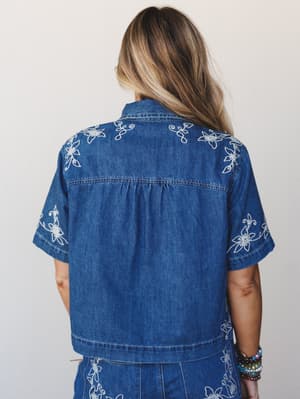 A back-facing shot of a model in a cropped short-sleeve denim shirt covered in white floral embroidery, paired with a matching embroidered denim skirt — the kind of set that makes a Saturday feel like something worth dressing for.