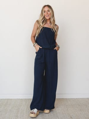 A navy strapless wide-leg jumpsuit with embroidered waist detail styled with espadrille platform sandals, beaded jewelry, and an easy smile — the kind of outfit that works as hard at brunch as it does at a summer concert.