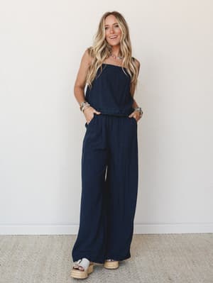 A navy strapless wide-leg jumpsuit with embroidered waist detail styled with espadrille platform sandals, beaded jewelry, and an easy smile — the kind of outfit that works as hard at brunch as it does at a summer concert.
