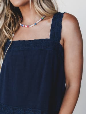 A close-up of a navy embroidered tank top with daisy-stitch detailing along the neckline and wide straps, styled with a playful mixed-bead and pearl necklace — the kind of effortless summer piece that looks equally good at brunch or on a boat.