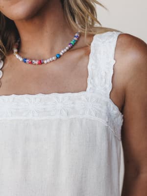 A close-up of a white linen tank top with tonal daisy embroidery trim along the square neckline and wide straps, styled with a colorful mixed bead necklace — easy, warm-weather dressing with a handmade feel.
