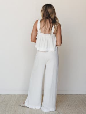 A back-facing shot of a white-on-white coord set — a cropped sleeveless top with lace trim and open back paired with flowy wide-leg pants, shot against a soft neutral wall with a relaxed, airy feel.