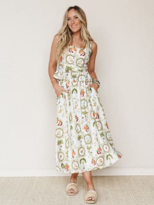 A smiling model in a breezy ivory midi skirt with a tropical tile print — palm trees, pineapples, and exotic birds framed in ornate medallions — styled with a matching sleeveless top and fringe slide sandals against a soft neutral studio wall.