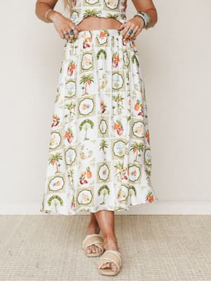 A flowy ivory midi skirt printed with framed tropical motifs — palm trees, pineapples, parrots, and botanicals — styled as a two-piece set with stacked rings, beaded bracelets, and raffia slide sandals for a warm-weather, free-spirited look.