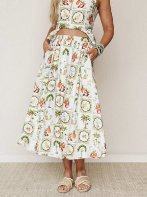 A flowy midi skirt in a vintage-tropical tile print — palm trees, pineapples, and fruit medallions on ivory — styled with a matching crop top and woven sandals for an effortlessly warm-weather feel.
