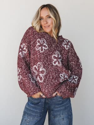 A relaxed, oversized burgundy marled knit sweater covered in loopy floral intarsia detailing, paired with wide-leg denim — the kind of sweater that makes a whole outfit feel effortlessly pulled together without trying.
