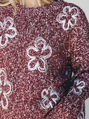 A close-up detail shot of a marled burgundy knit sweater covered in raised cream floral appliqués — the kind of sweater that makes a whole week feel warmer.