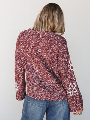 A back-view shot of a burgundy marled knit sweater with white floral embroidery along the sleeves, paired with relaxed medium-wash jeans — cozy and personal, like something you'd reach for every single weekend.