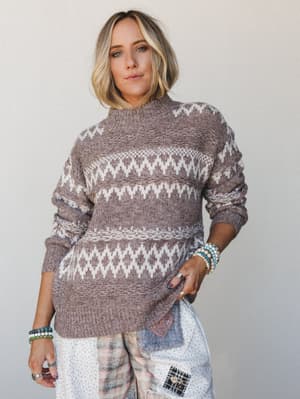 A relaxed fair isle knit sweater in warm mocha and cream, styled with patchwork wide-leg pants and stacked beaded bracelets — the kind of outfit that feels as good as it looks on a slow Saturday.