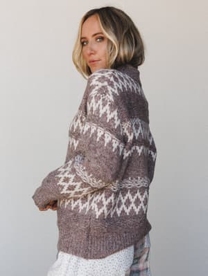 A model shot from behind and over the shoulder, looking back at the camera with an easy confidence — the sweater's fair isle knit pattern in mocha and cream does all the talking.
