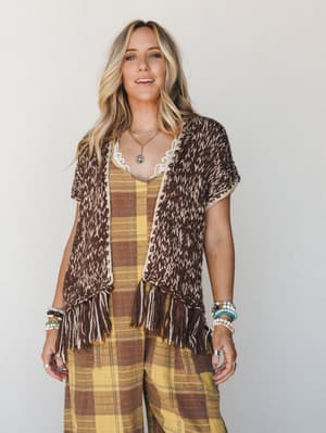 A layered boho look built around a chunky knit fringe vest in mocha and cream, worn open over a golden yellow plaid wide-leg set — the kind of outfit that looks like it was pulled together effortlessly but absolutely wasn't.