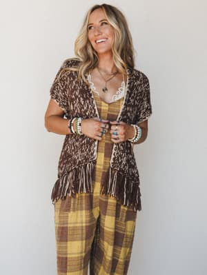 A smiling model layers a fringed open-front knit vest in mocha and cream over a yellow plaid wide-leg jumpsuit, styled with stacked beaded bracelets and layered gold necklaces — the whole look reads like a warm autumn afternoon with nowhere to be and