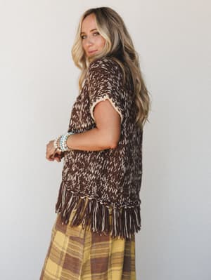 A model shot from the back at a three-quarter turn, wearing a chunky open-knit fringe sweater vest in mocha and cream over a yellow plaid maxi skirt, styled with turquoise beaded bracelets — earthy, free-spirited, and effortlessly put-together.