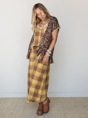 A plaid maxi dress layered under a fringed knit vest in mocha and cream, styled with turquoise stacked bracelets and brown clogs — earthy, effortless, and very much autumn in the best way.