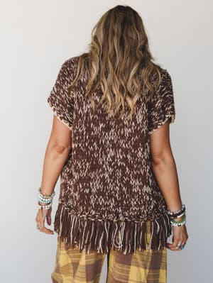 A back-view shot of a chunky knit fringe vest in mocha and cream marled yarn, styled over plaid wide-leg pants with stacks of beaded bracelets — earthy, relaxed, and very much the outfit you wear when you actually want to enjoy your weekend.