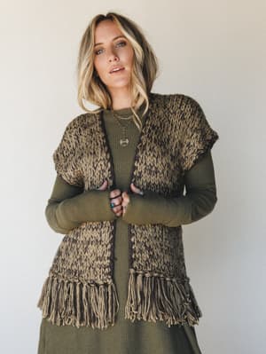 A chunky hand-knit fringe vest in mixed olive and dark brown tones layered over a long-sleeve ribbed olive dress — earthy, textural, and the kind of outfit that makes a Tuesday feel intentional.