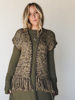 A chunky hand-knit fringe vest layered over an olive ribbed maxi dress, styled with layered gold necklaces — earthy, relaxed, and the kind of outfit that looks like it was thrown on effortlessly but absolutely wasn't.
