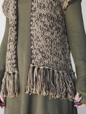 A close-up torso shot of a chunky hand-knit fringe vest layered over a ribbed olive green long-sleeve dress, styled with silver rings and colorful nails — earthy, textural, and very much the kind of outfit you'd wear on purpose.