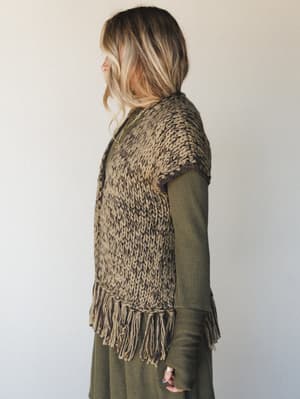 A side profile shot of a chunky knit fringe vest in mixed olive and brown tones layered over a long-sleeve olive ribbed dress — earthy, textural, and the kind of outfit that feels like it has a whole personality.