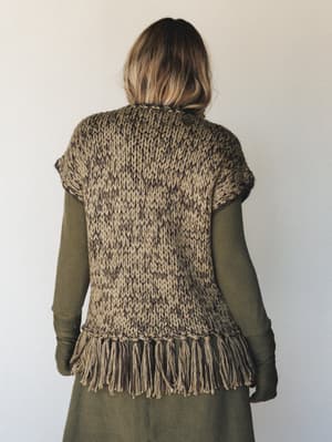 Shot from the back, a chunky knit fringe vest in a warm tan-and-dark-olive marled mix layers over an olive long-sleeve midi dress — the kind of outfit that feels intentional without trying too hard.