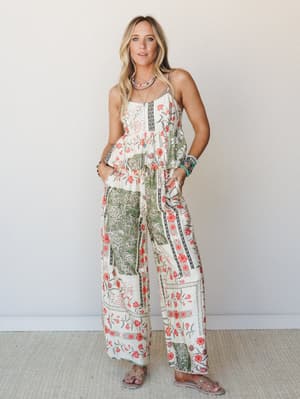 A flowy floral patchwork set — wide-leg pants and a smocked spaghetti strap top in cream, coral, and sage — styled with layered jewelry and flat sandals against a soft neutral studio wall, giving off effortless warm-weather energy.