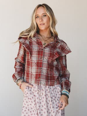 A ruffle-trimmed plaid blouse in brick red is layered over a soft floral skirt — the kind of effortless mix that feels intentional without trying too hard, finished with stacked beaded bracelets and a layered gold necklace.