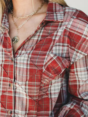 A close-up of a brick red plaid flannel button-down with a standout quilted ruffle detail at the chest, styled with layered gold chain necklaces for an effortlessly personal touch.