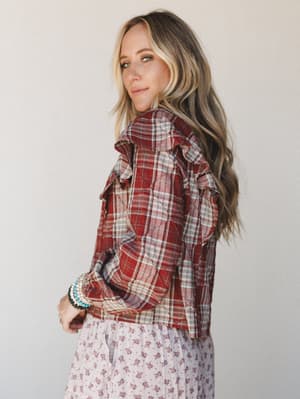 A model shot over the shoulder in a brick red plaid ruffle-trim blouse layered over a soft floral skirt — the kind of effortless mix-and-match that feels intentional without trying too hard.