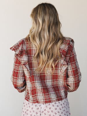 Shot from behind, this quilted plaid jacket in brick red layers over a floral midi skirt — the ruffle shoulder trim and diamond quilting give it that sweet mix of cozy and considered that's hard to pull off but easy to wear.