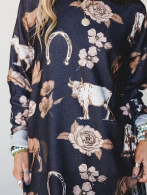 A close-up torso shot of a dark navy sweater covered in a whimsical western-meets-romantic print — horses, cattle, horseshoes, and roses all living their best life together — styled with layered beaded bracelets and a gold coin necklace.