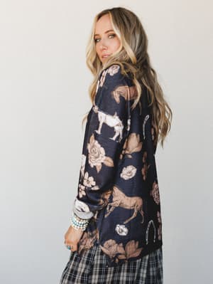 A model shot over the shoulder in a navy graphic sweater printed with horses, horseshoes, and roses — layered over a plaid skirt and stacked with beaded bracelets, giving off that effortless Western-boho energy.