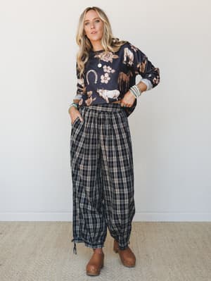 A relaxed, pattern-mixed outfit — a dark graphic sweatshirt printed with horses, florals, and horseshoes layered over voluminous plaid pants, finished with tan clogs and turquoise bracelet stacks for that effortless western-boho feel.