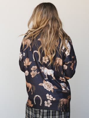 Shot from behind, this navy sweater does all the talking — a whimsical western-floral print featuring horses, longhorn cattle, horseshoes, and blooming roses layered over a dark ground, paired with a plaid skirt peeking out at the hem.